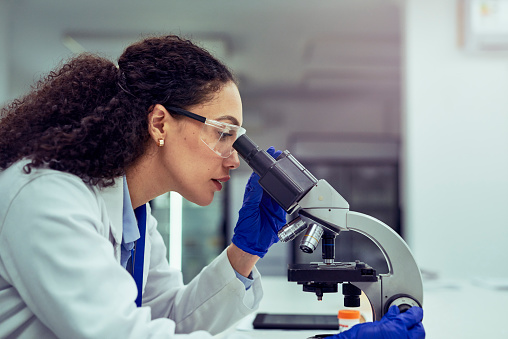 Female scientist wearing safety goggles and gloves, focusing on analyzing test sample under microscope in laboratory