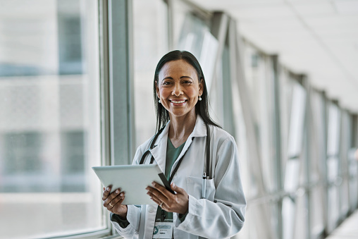 Female doctor holding digital tablet, smiling at camera beside window at corridor in hospital