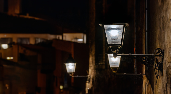 Antique wrought iron street lamps lit and anchored to a stone wall of an old medieval house at night with more old houses at night