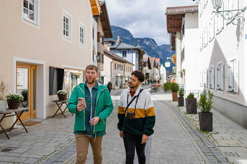 A medium front view of a male couple who are exploring in Garmisch town centre which is a little Bavarian town in the European alps in Germany. They are admiring the local sights and scenery and navigating their way around the area using their mobile phones map.