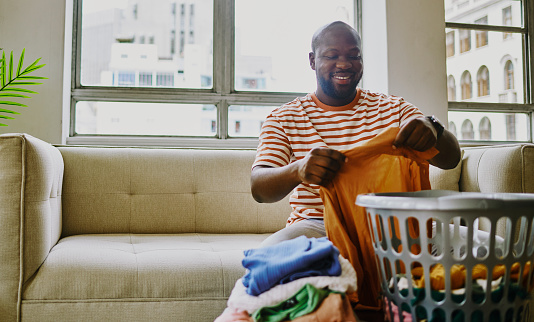 A young black man sitting on his sofa in his living room while folding his laundry. Stock photo, copy space