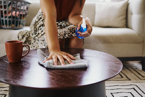 A shot of an unrecognized woman sitting while spraying and wiping her table in her living room. Stock photo