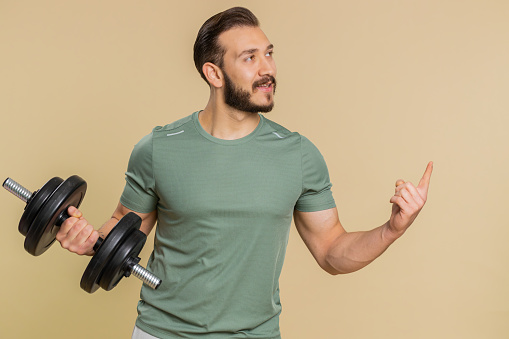 Athlete man showing pointing empty place, advertising area for commercial text, copy space for goods promotion, recommendation. Middle eastern sportsman guy weightlifting dumbbell on beige background
