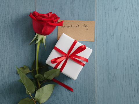 three red roses with gift on blue wood table with valentines day paper card shot from above