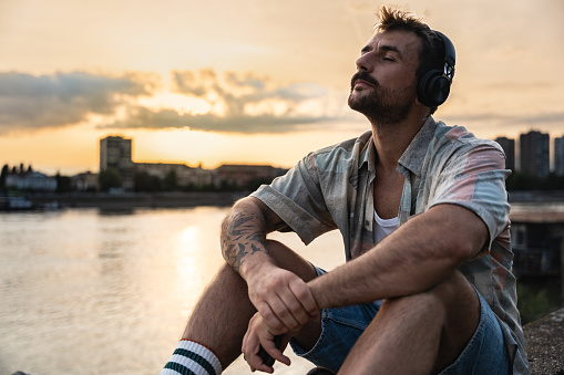 Happy teenage male tourist sitting on sea promenade of an old city in sunset. Young man world traveler singing his favorite song on headset, making a break from walking through the city.