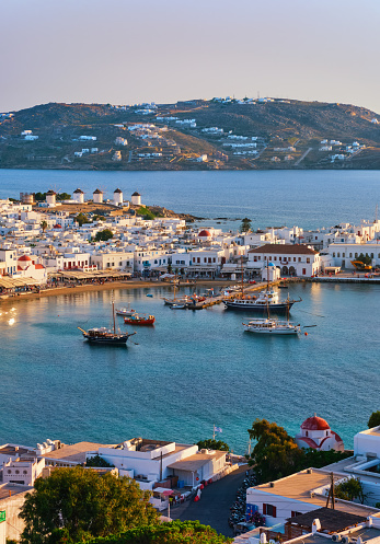 Beautiful sunset view of Chora, main town of Mykonos, Cyclades, Greece and its harbor and port. Golden hour, harbor, cruises, ship, whitewashed houses and distant islands in hazy background. Vacations, leisure, Mediterranean lifestyle. Vertical shot
