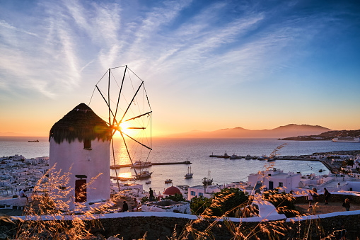 Famous traditional white windmill overlook civil port and harbor of Mykonos, Cyclades, Greece at sunset sky. Beautiful sky, sun touch sea horizon, Chora, main town of island, in evening lights and shadows.