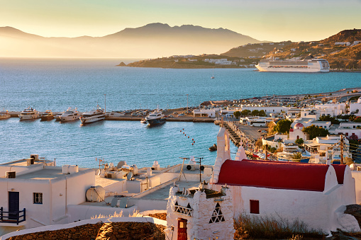 Beautiful view of mediterranean island town, Mykonos, Greece at sunset. Port, harbor, boats, sailboats, yachts, cruise liner moored by jetty. Famous whitewashed houses, white church with red roof, hazy horizon. Summer vacations, romantic weekend
