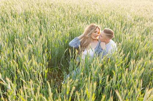 Young happy married couple is sitting in tall green grass in meadow on sunny day,hugging and looking at each other. Waiting for baby. Wonderful moments. Family relations. Positive emotions.