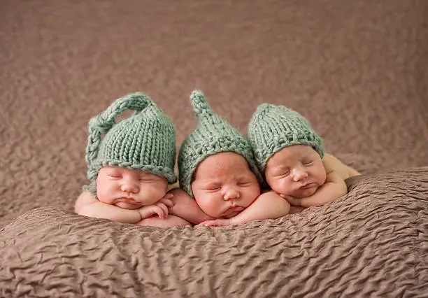 Identical triplet girls sleeping peacefully on a soft blanket; wearing knit hats.