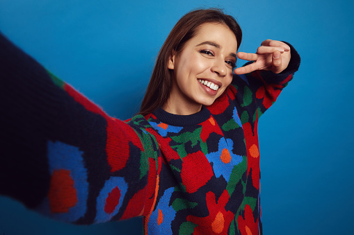 Close up portrait of excited female in colorful sweater making peace gesture over eye and smiling while making selfie, isolated over blue background.