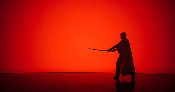 Athletic Man Practicing Kendo Martial Arts with a Katana or a Bamboo Sword. Male Does Cosplay, Swinging the Cold Weapon in a Modern Dojo Style Digital Space with a Red Backdrop on a Screen