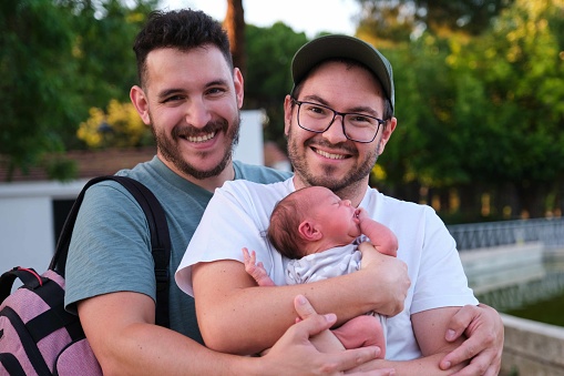 Gay couple with their newborn baby in a park smiling and looking at camera. Four days old infant, parenthood concept.