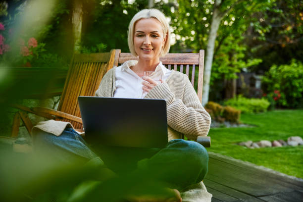 Adult woman sitting relaxed with laptop on garden terrace during summertime stock photo