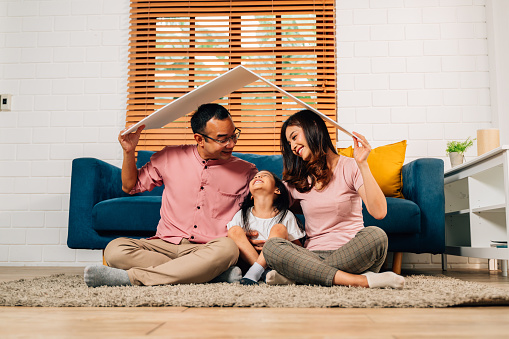 Happy family holding symbolic cardboard roof of life insurance and sitting on couch at home, Family care, Protection and safety life insurance concept, Mortgage loan and investment security for future