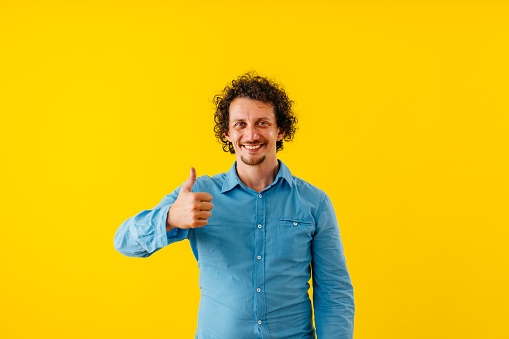Portrait of a handsome young man with thumbs up standing in front of a bright yellow wall.