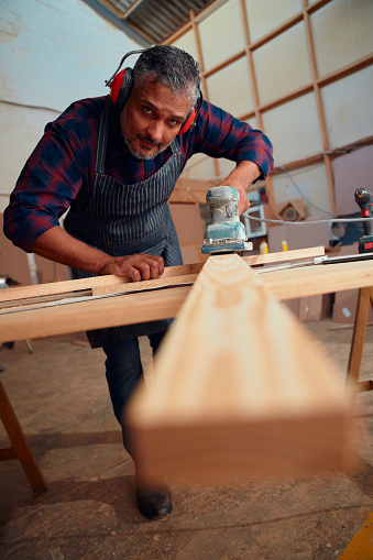 Multiracial mid adult man wearing ear muffs using power tool on wood plank in woodworking factory