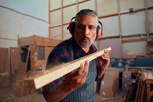 Multiracial mid adult man wearing ear muffs blowing on wood plank in woodworking factory