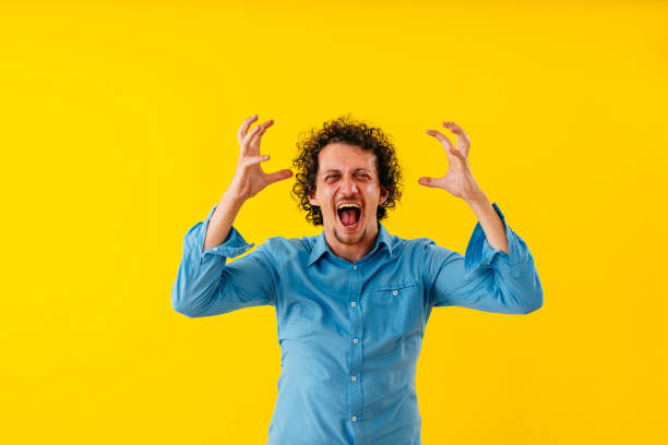 Portrait of an angry young man standing in front of a yellow wall and shouting.