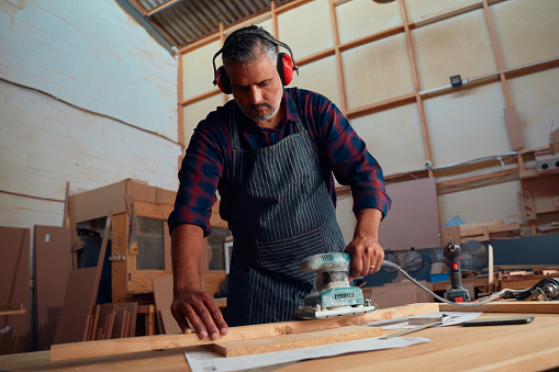 Multiracial mid adult man wearing ear muffs and using power tool on wood plank in woodworking factory