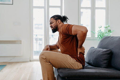 A young African American man is sitting in his living room and leaning forward from his back pain.