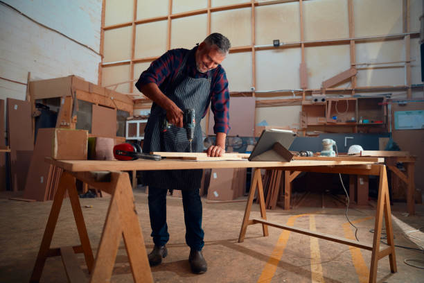 istock Mid adult man smiling while using drill on timber next to digital tablet on table in woodworking factory 1530778987