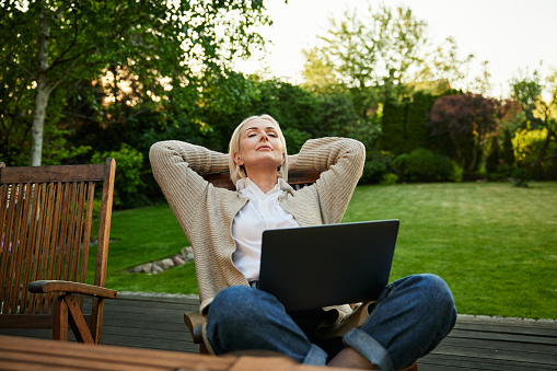 Mature woman with eyes closed relaxing on backyard garden terrace sitting on a chair with laptop
