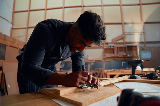 Multiracial young man writing with pencil while using tool on wood at woodworking factory