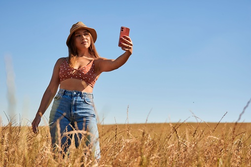 A Caucasian female in a straw hat taking a selfie with her phone in a wheat field