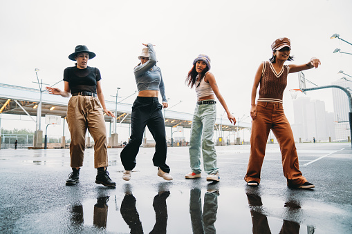 Four female friends are dancing together near a puddle in a basketball court. They are wearing street style clothes.