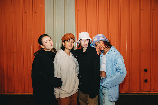 Portrait of four friends wearing cool street style clothes looking at camera against an orange wall. They are wearing hats. Hip hop dancers.