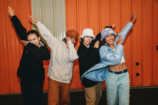 Portrait of four friends wearing cool street style clothes making dabbing movement against an orange wall. They are wearing hats. Hip hop dancers.