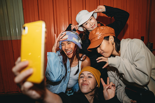 Young adult women wearing cool street style clothes are taking a selfie at night. They are wearing hats. Hip hop dancers.