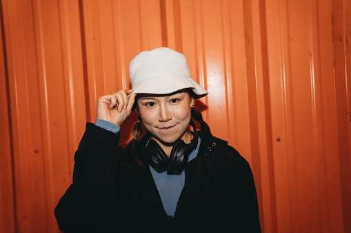 Portrait of a young adult woman looking at camera against an orange wall in the city. She's wearing cool street style clothes and a hat.