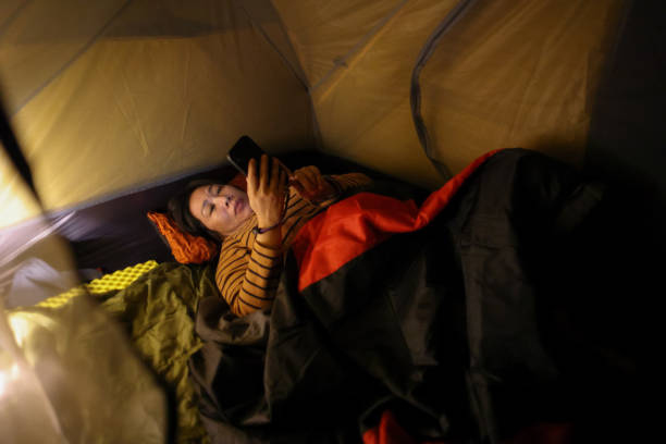 The girl is lying in a sleeping bag on a warm mat near a tent outdoor. Family camping recreation, hiking equipment.