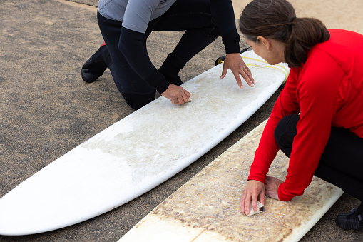 A close-up of a senior couple waxing their surfboards for traction at the beach before going surfing. they are wearing wetsuits and coloured rash vests. They are surfing at Tynemouth beach on the North East coast of England.