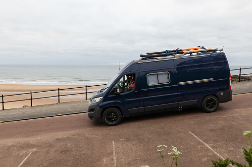 A wide-angle view of a mature couple parking up at the beach in their campervan. they are assessing the surf conditions before going surfing. There are inflatable paddleboards on top of the van roof. It is an overcast day.