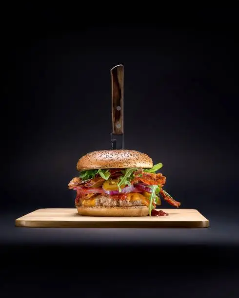 A vertical shot of a burger on a wooden cutting board, with a sharp knife protruding from the top