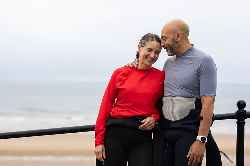 A mature surfing couple embracing each other at Tynemouth Beach in the North East of England. The gentleman has his arm around his partner as he whispers into her ear. They are both portraying happy emotions and bright smiles. They are both wearing wetsuits around their waist with coloured surfing rash vests.