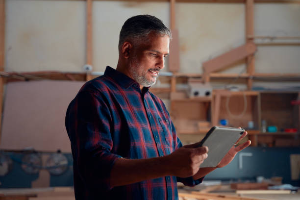 istock Mid adult man in checked shirt smiling while using digital tablet at woodworking factory 1530859622