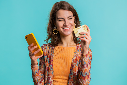 Sincere young woman customer using credit bank card and smartphone while transferring money, purchases online shopping, payment. Finance and internet. Pretty girl isolated alone on blue background