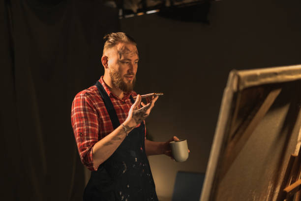 istock Male artist standing in his modern creative studio with large canvas, holds phone talks on speakerphone with friend, makes voice recognition or request uses virtual assistant 1530888922