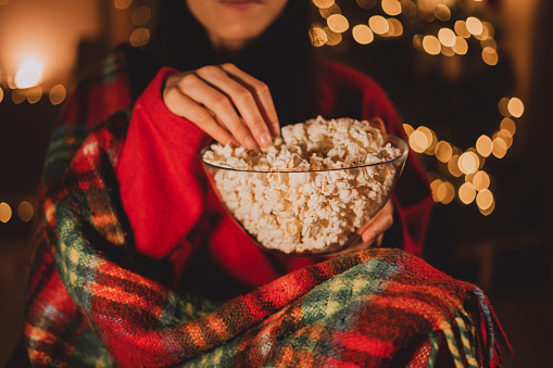 Cheerful woman eating popcorn and watches christmas comedy movie on cable TV at home in evening alone, close-up. Christmas traditions concept