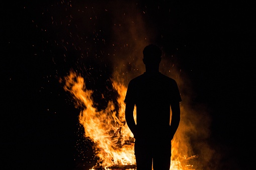 A silhouette of a person standing against a backdrop of a burning bonfire