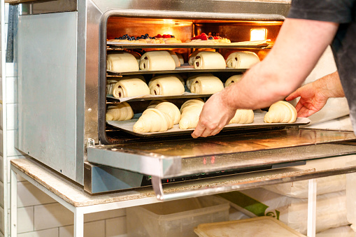 In this close-up photo, an anonymous bakery worker deftly places trays of croissants and chocolate pastries into the oven, creating a mouthwatering display of baking expertise.