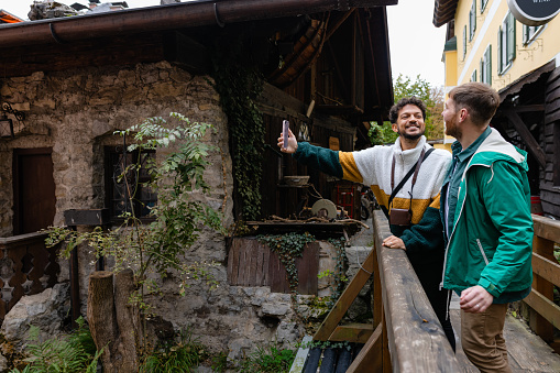 A side view of a gay couple who are exploring in Garmisch town centre in southern Germany.  They are impressed with the beautiful traditional German architecture. They are walking over a wooden footbridge through the little alleys and stopping to take a selfie.