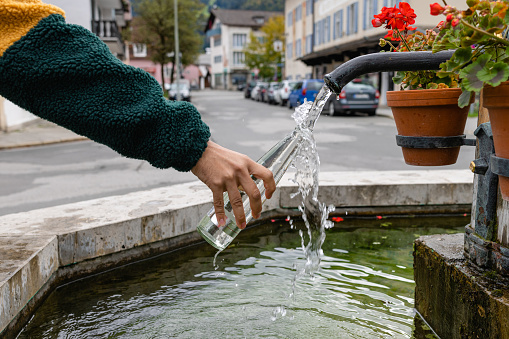 A close up of an unrecognisable man in Garmisch in south Germany. He has stopped at a water fountain which has Geranium plants around the tap in the town centre which provides the town with flowing free spring water from the mountains, He is filling up his reusable glass bottle.