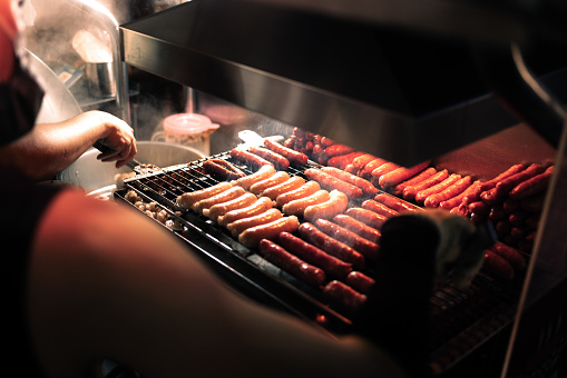 A close-up shot of a night market vendor grilling sausages. The sausages are enveloped in a cloud of smoke under the warm glow of the lights, making them look irresistibly delicious.
