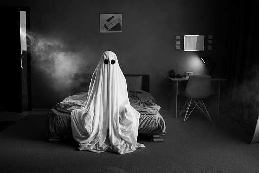 Ghost in a white sheet sits on a bed in bedroom in smoke, black and white photo. Halloween concept. Copy space