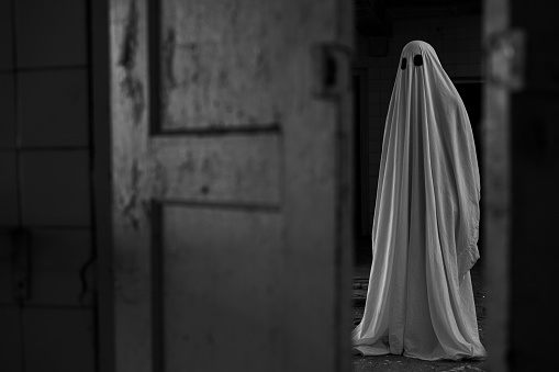 Scary ghost covering in white sheet on dark background near open door in abandoned build. Black and white photo. Copy space